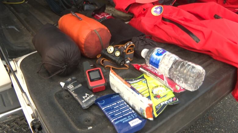 A pick up truck tailgate is loaded with energy bars, a water bottle, flashlight, GPS, sleeping bag, emergency blanket and warm clothing.