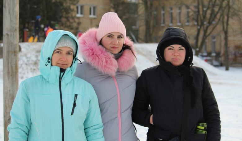 Three women wearing winter coats stand outside in the snow.