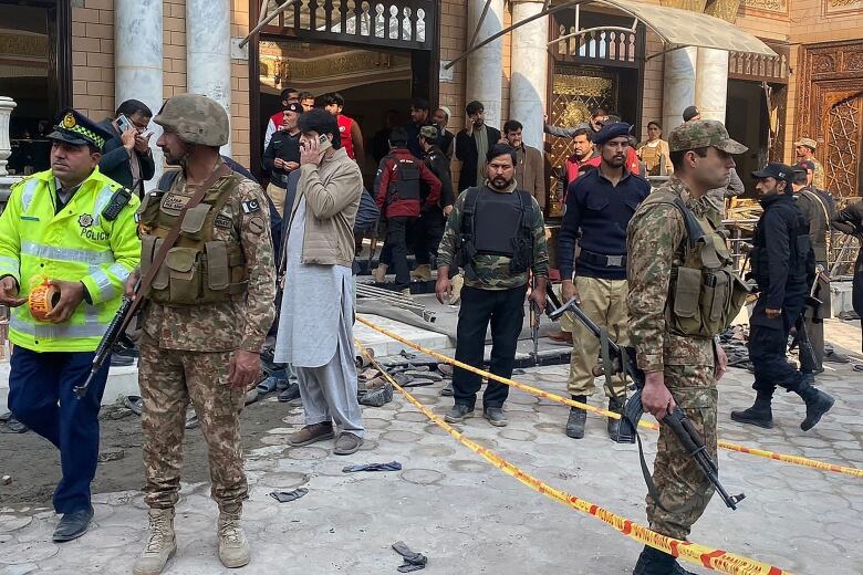 Men in fatigues with weapons, police officers and civilians are shown standing in an outdoor scene.