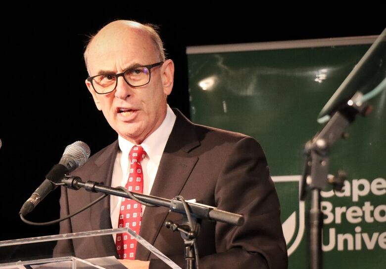 A balding man with glasses, a dark suit, white shirt and red tie speaks at a microphone.