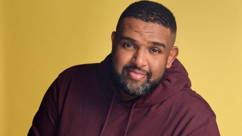 A man with short hair standing in front of a yellow wall wearing a maroon hoodie.