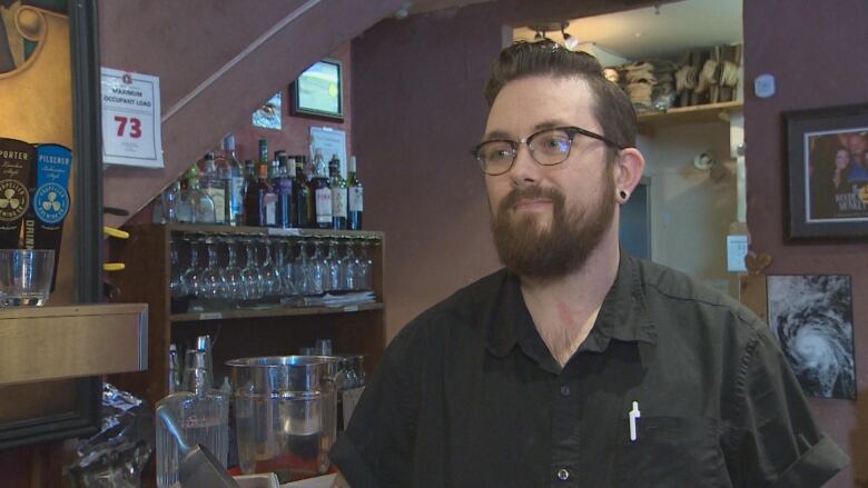 A man wearing glasses and a black shirt stands in front of a bar.