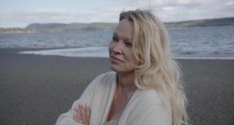 Woman in blonde hair and white coat stands on beach in front of ocean waves