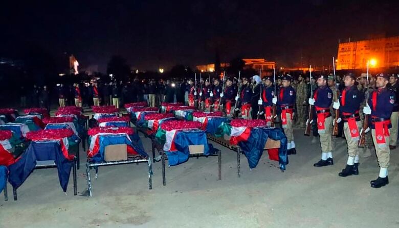 Armed soldiers stand at attention beside rows of coffins draped in flags.