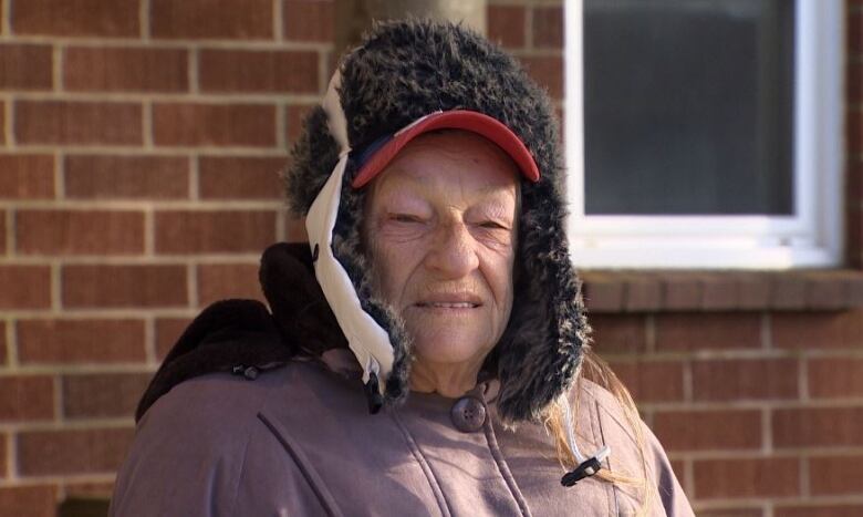 An elderly woman wearing a purple jacket and a black hat sitting in front of a brick wall. 