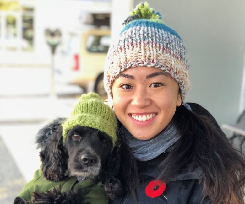 A woman in a colourful toque and black coat, with a red poppy, holds a dog in a green sweater and toque.