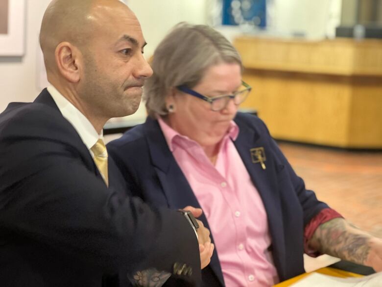 Man in suit holding hand of woman in suit, pink shirt.