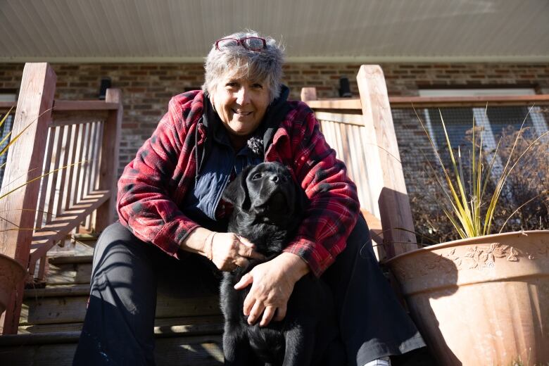 A woman wearing a red plaid jacket sits on steps of a house while hugging a Labrador retriever.