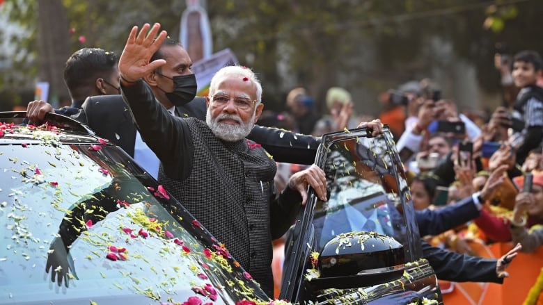 Indian Prime Minister Narendra Modi exits a vehicle and waves to supporters. There are two security officers behind him and there is a crowd in the background. The car is covered in flower petals. 