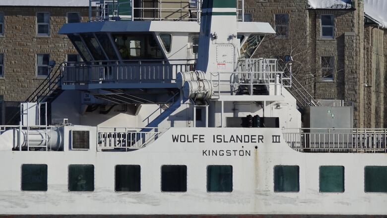 The words Wolfe Islander III are written in big green letters on the side of a white ferry. It's a close up photo of the name. A grey brick building is in the background.