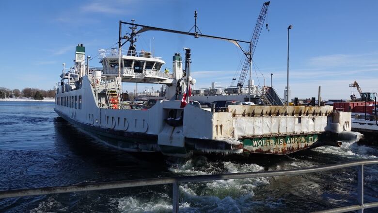 Icicles hang off the back of a ferry and the water bubbles up as it powers past a dock covered in construction equiptment.