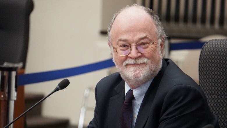 A seated man with white-grey hair and facial hair, wearing a suit, smiles at the camera from behind a microphone.