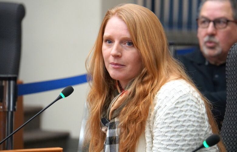 A woman with long red hair and blue eyes sits behind a microphone.