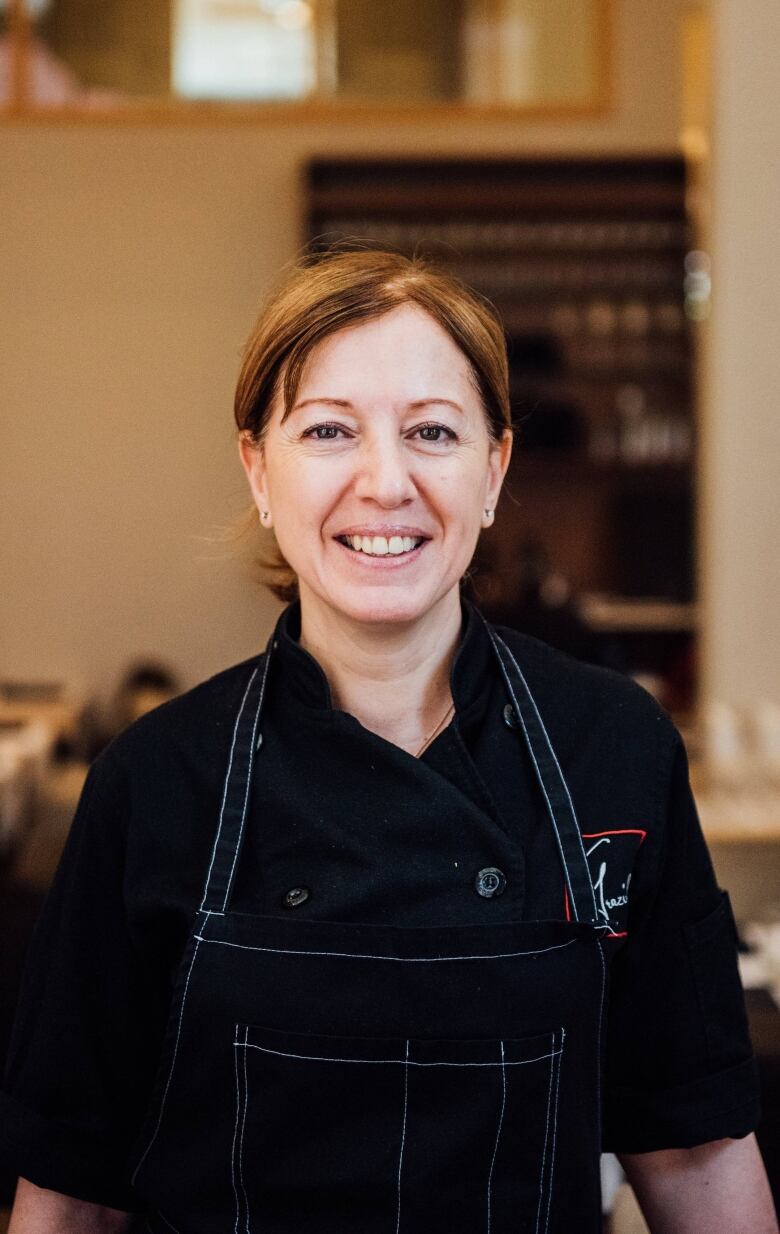 Woman in black chef's jacket and black apron. Restaurant tables in background.