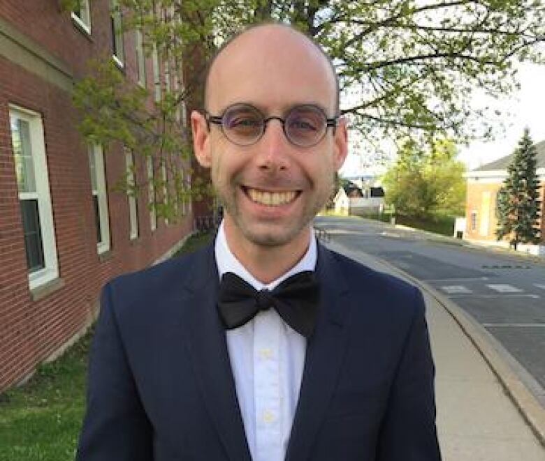 A man in a black suit and bowtie stands on a sidewalk outside smiling.