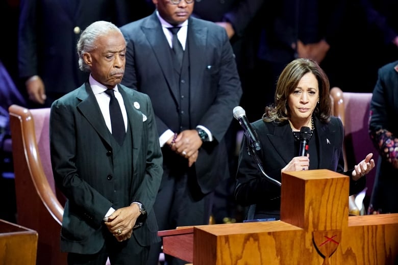 A man and a woman in dark suits stand near a large wooden cross. The man looks down with his hands clasped. The woman speaks into a hand-held microphone.