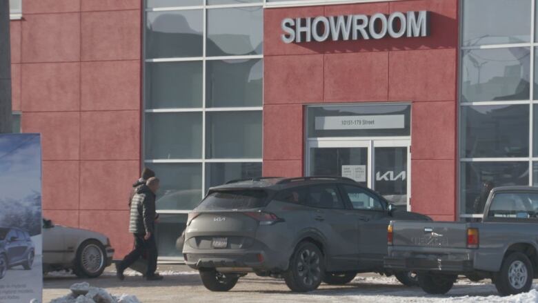 Two men  a KIA salesman and Go Public's secret shopper  enter KIA West Edmonton car dealership. The exterior of the dealership is red. Both men are wearing coats. 