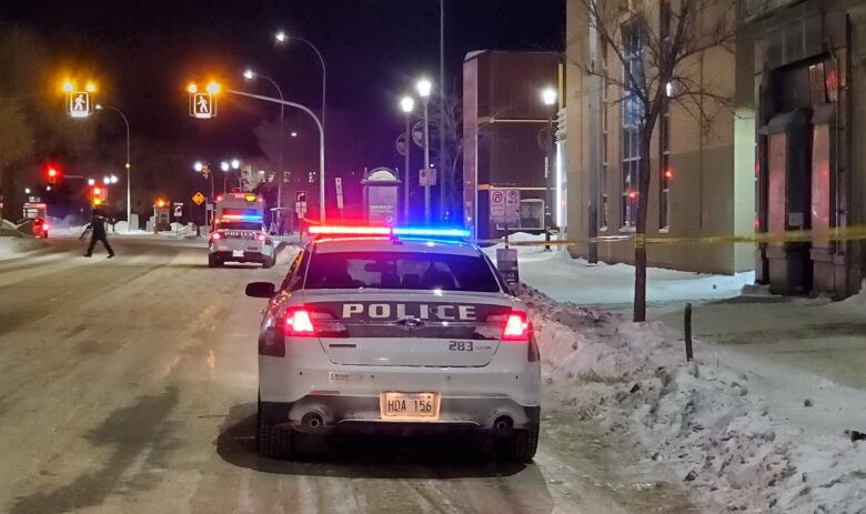 A police cruiser, with its red and blue lights on, is parked next to a snowy curb. Yellow police tape can be seen to the right of the car, across the sidewalk.