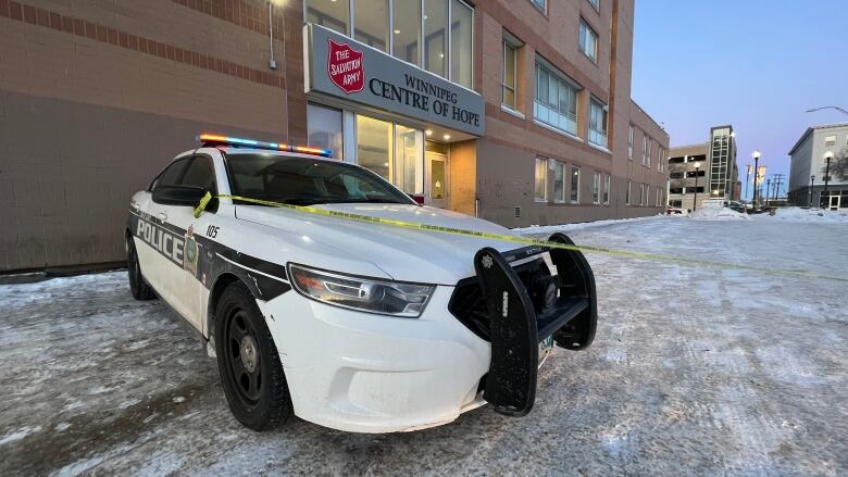 A police car with yellow caution tape tied to its side mirror, blocks the snowy parking lot of a building. A name above the door to the building says Winnipeg Centre of Hope.