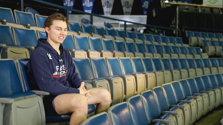 A teenaged boy sits with blonde hair, wearing a navy blue hoodie and shorts, sits in a bleacher seat at an empty hockey arena.