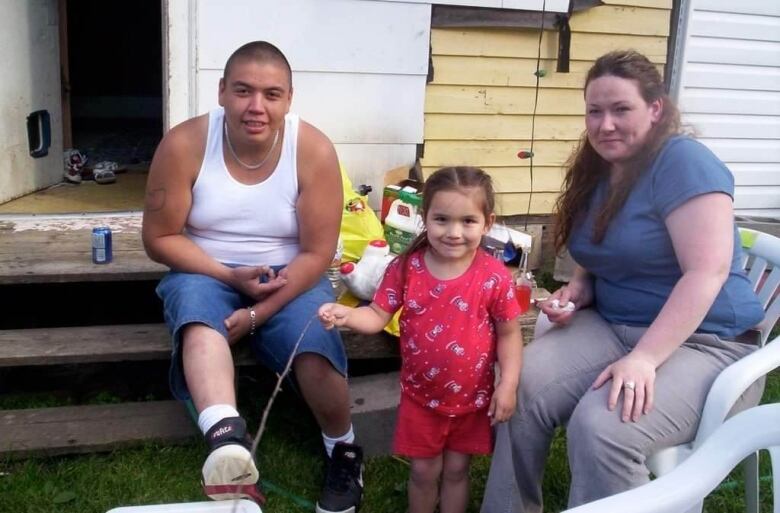 A man and woman are seated in a backyard with a toddler standing between them.