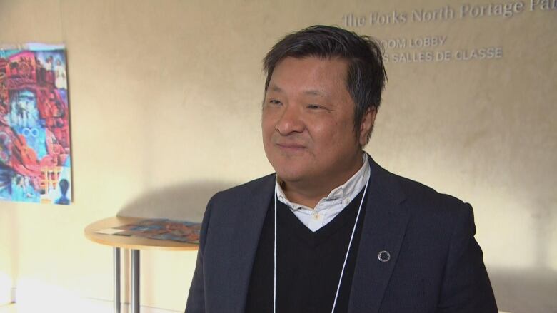 A man in a dark blazer and sweater speaks to a reporter.