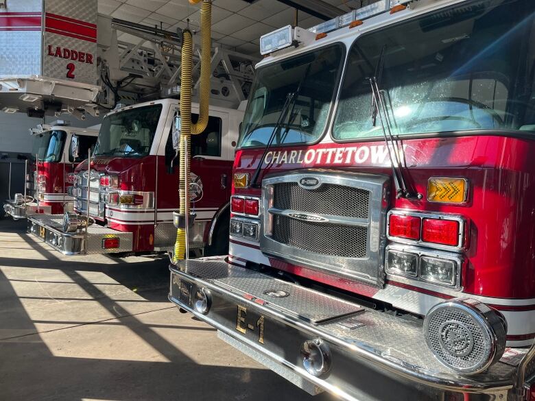 Three red fire trucks sit in the bay of a downtown fire hall, with the sun shining on the trucks. 