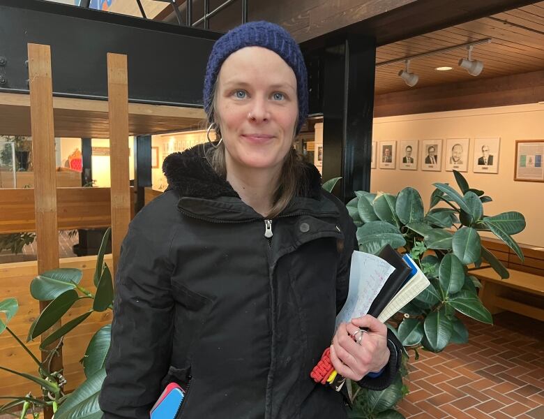 A woman in a hat and winter coat stands inside a building, holding some books and papers.