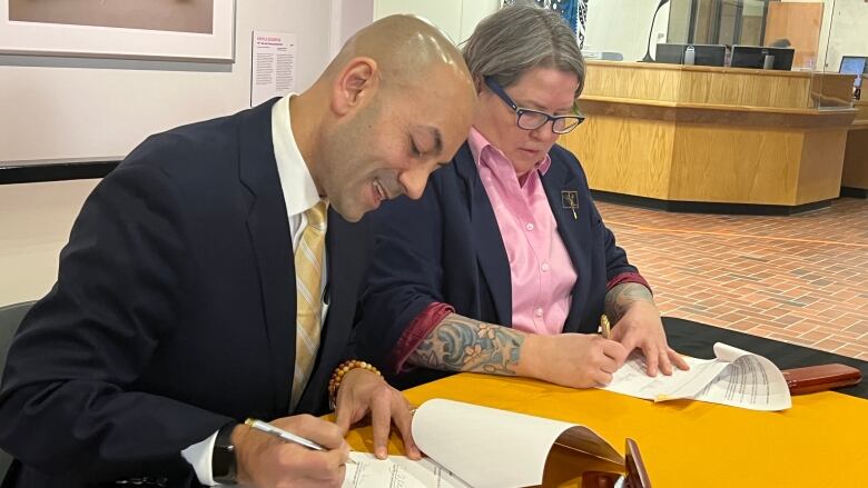 A man in a suit and a woman in a blazer and pink button up sign documents at a table. 