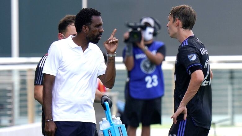 A coach in a white polo points his finger as he talks to a soccer player on the sidelines.