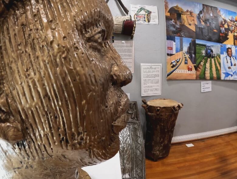 A close up shot of the side profile of a brass head bust. Behind it, and slightly out of focus is an African drum and a large painting. 