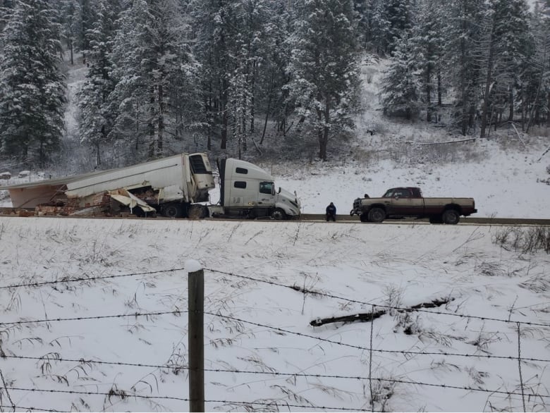 The trailer of a transport truck is smashed and its contents are lying on the highway. A pickup truck faces it, a man sits on the side of the highway.