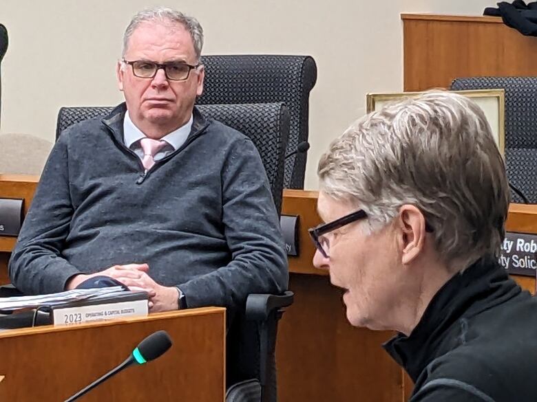 A side shot of Thunder Bay resident Karen Rooney speaking to city council while sitting at a desk in the council chambers in front of a desk-top microphone.  City manager Norm Gale can be seen in the background looking on with an earnest expression on his face.