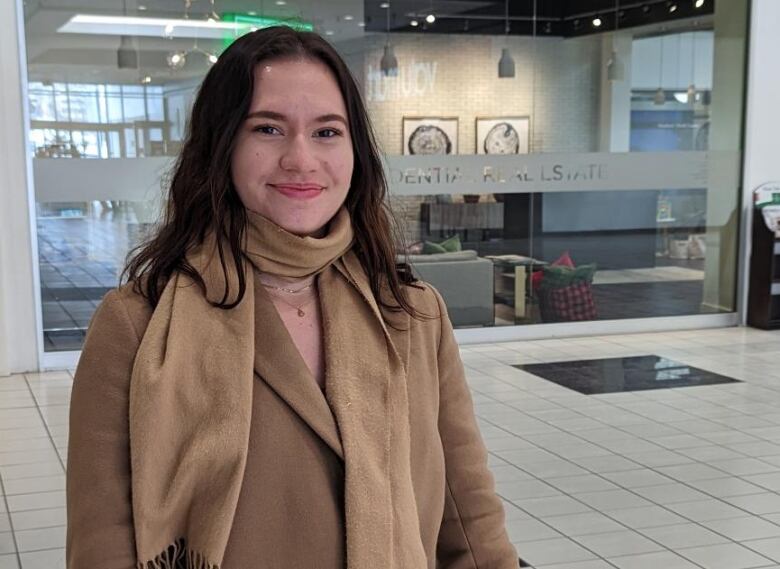 Student in a camel coloured jacket and scarf stands in front of a storefront smiling.