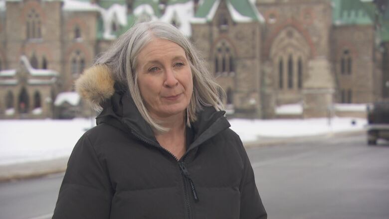 A grey haired woman wearing a black winter jacket with a fur trimmed hood standing outside on a street in the winter. 