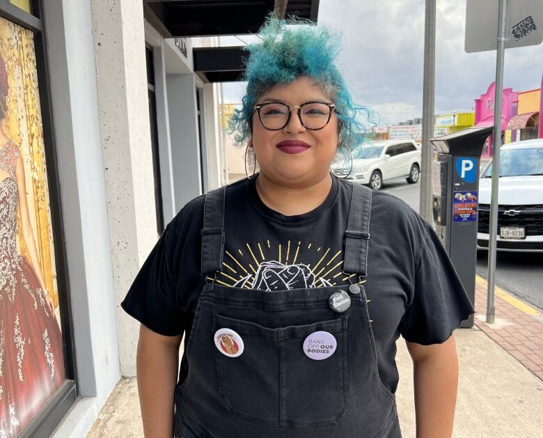 A woman with blue-green dyed hair wearing glasses and coveralls stands on a sidewalk.