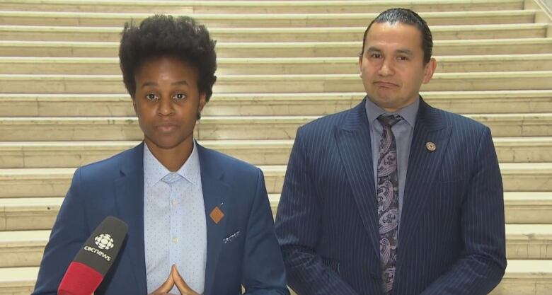 Two people stand behind a podium and face the camera. One, at left, wears a blue collared shirt and blue suit jacket and has black hair. The one at right is in a blue pin-striped suit, a collared shirt and tie, with long black hair pulled back.