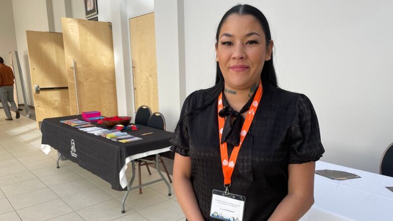 An Indigenous woman stands next to a table at a conference, holding tobacco and wearing a traditional skirt.