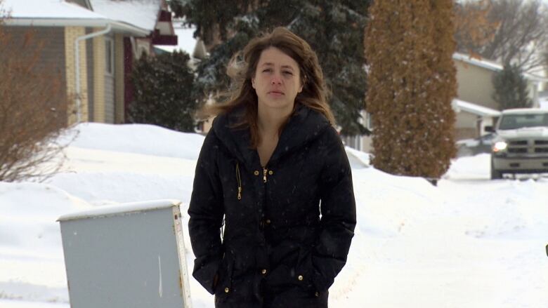 A white woman with long brown hair and a black parka is shown from the waist up, walking down a snowy suburban street.