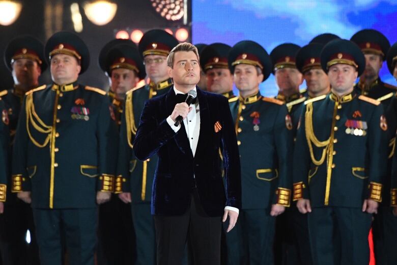 A blond-haired man, in a navy tuxedo jacket, white shirt and black bowtie, stands with a microphone in front of lines of men in green and gold military uniforms.