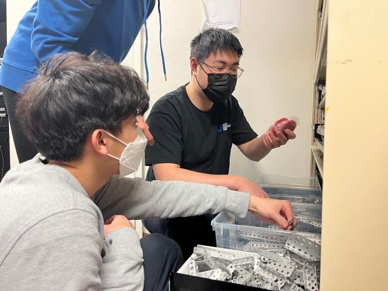 two young people wearing masks crouch over a bucket of parts. one of the boys is wearing a black shirt and looks like he's explaining 