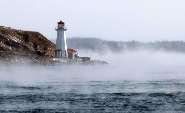 A view of a lighthouse on an island across the water. Mist rises from the water.