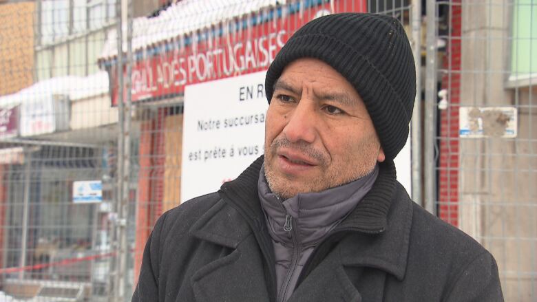 A man standing in front of a charred and boarded up grocery store wearing a black jacket and black tuque. 