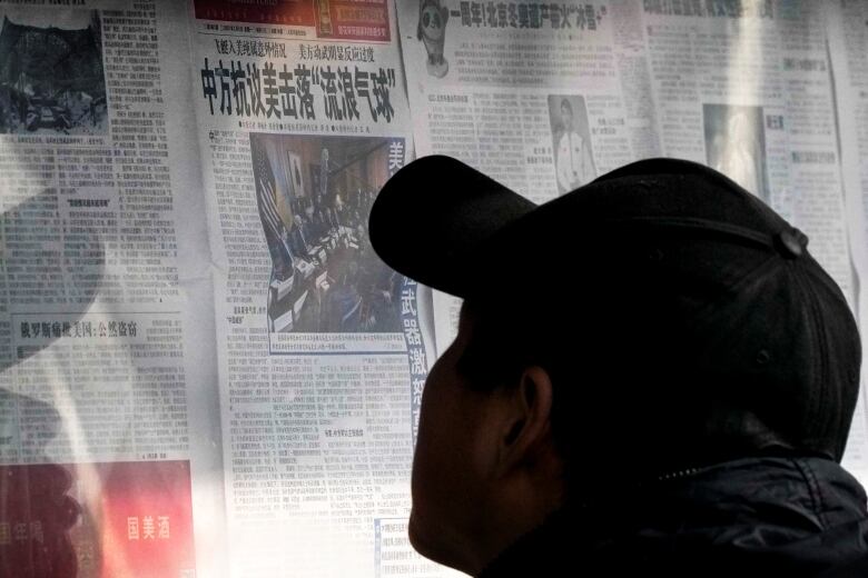 A man in a baseball cap reads a newspaper posted up at a newstand in Beijing, China.