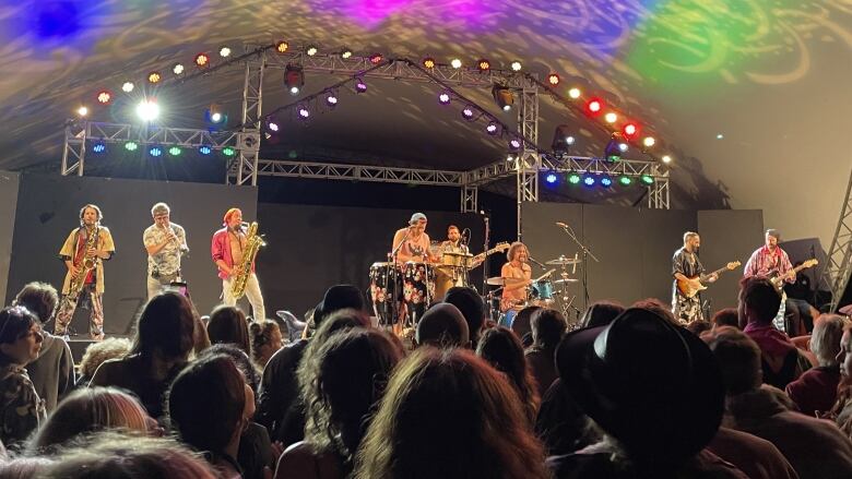 A crowd stands in front of a colourful, lighted stage where a big band with several brass instruments, three guitars, drums and percussion are performing.