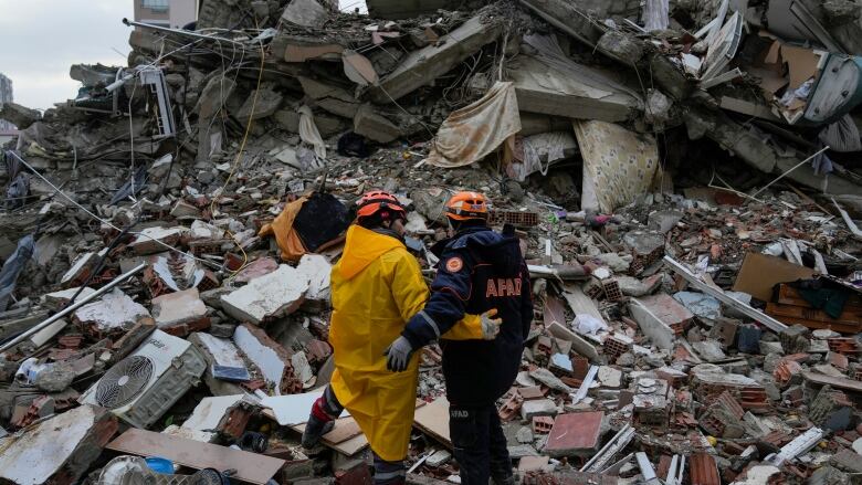 Two rescue workers stand with their backs to the camera on top of a pile of rubble, their arms around each other.