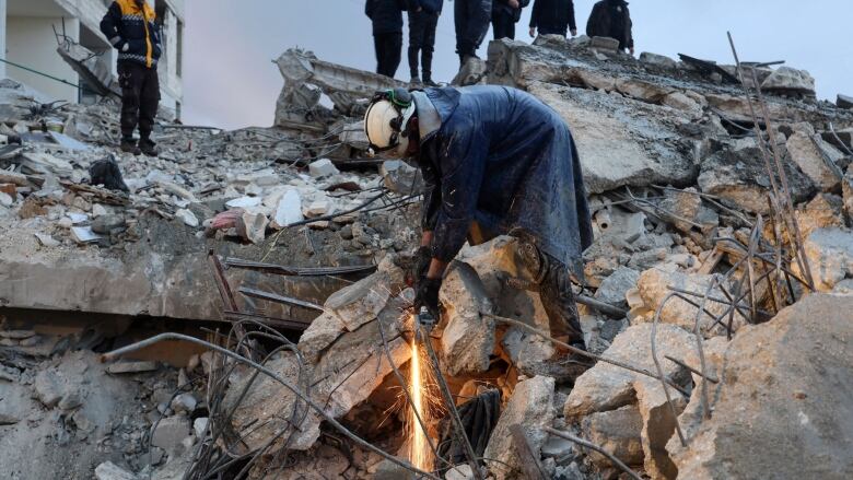 A man with a white helmet cuts through concrete as other people stand above, on a pile of rubble, watching. 