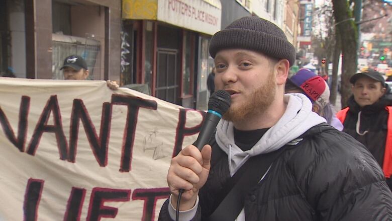 A man in a black jacket and black toque speaks into a microphone at a protest in Vancouver. 