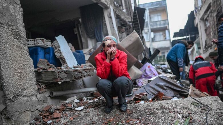 A woman appears to cry with hands held to her face while crouching on rubble in an urban setting.