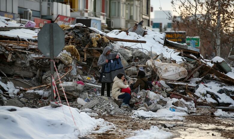 A man stands on a pile of snow-covered debris, holding a jacket, while a woman is sitting in front of him and holding a child.